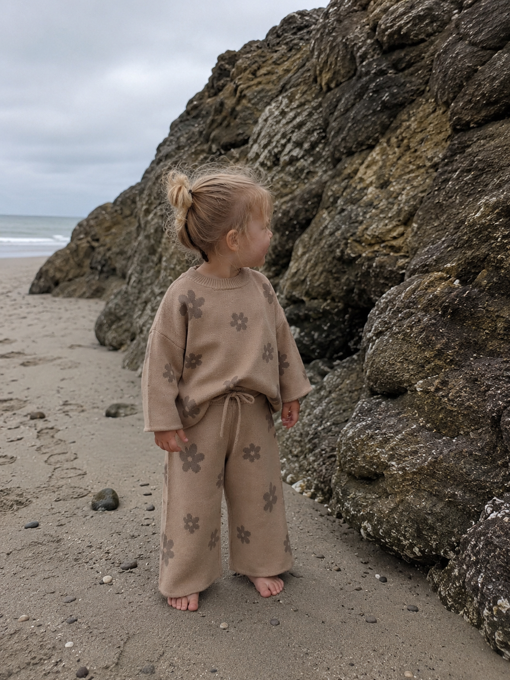 Child in a beige outfit with floral patterns standing on a rocky beach.